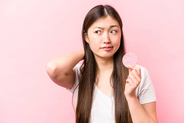 年轻的亚洲女人 手里拿着用粉红背景隔离起来的面部海绵 后仰着头 思考着 做出选择 — 图库照片