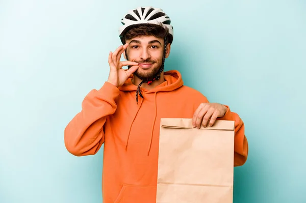 Young hispanic man wearing a helmet bike holding a take away food isolated on blue background with fingers on lips keeping a secret.
