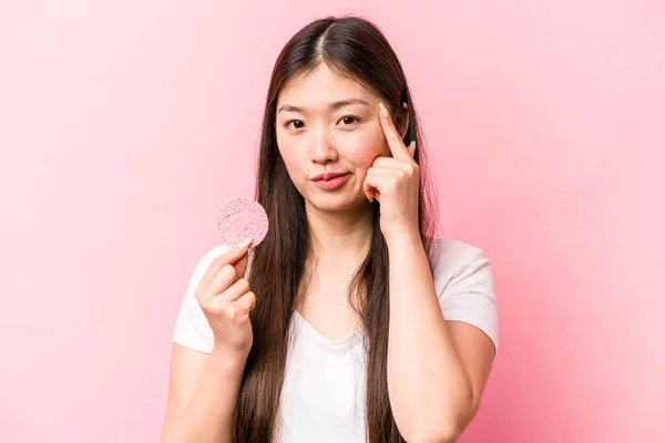 年轻的亚洲女人拿着用粉红背景隔开的面部海绵 用手指 专注于一项任务 — 图库照片
