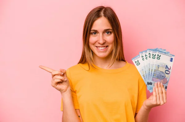 ピンクの背景に銀行券を持っている若い白人女性は笑顔と脇を指して 空白のスペースで何かを示しています — ストック写真