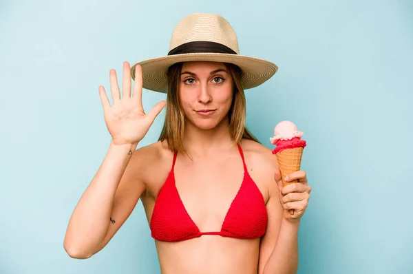 年轻的高加索女人穿着比基尼 手里拿着一只蓝色背景的冰淇淋 高兴地微笑着 用手指展示着第五位 — 图库照片