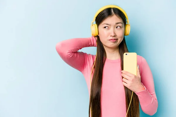 Young asian woman listening to music isolated on blue background touching back of head, thinking and making a choice.