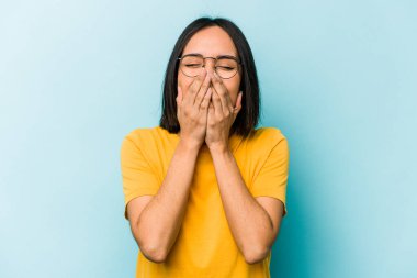 Mavi arka planda izole edilmiş genç İspanyol kadın bir şeye gülüyor, elleriyle ağzını kapatıyor..