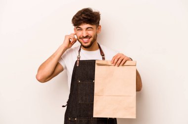 Beyaz arka planda izole edilmiş, kulakları elleriyle kapatan bir paket taşıyan genç İspanyol tezgahtar..