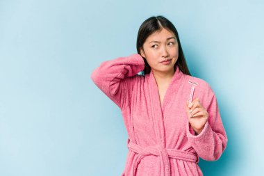 Elinde jilet tutan genç Asyalı kadın mavi arka planda izole edilmiş kafasının arkasına dokunuyor, düşünüyor ve bir seçim yapıyor..