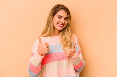 Beyaz tenli genç bir kadın bej arka planda izole edilmiş. Baş parmağını kaldırıyor, gülümsüyor ve kendinden emin..