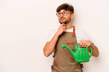 Genç bahçıvan, elinde su dolu bir teneke kutu tutan, şüpheli ve şüpheci bir ifadeyle beyaz arka plana bakan bir İspanyol..