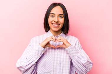 Pembe arka planda izole edilmiş genç İspanyol kadın gülümsüyor ve elleriyle kalp şeklini gösteriyor..