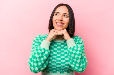 Pembe arka planda izole edilmiş genç İspanyol kadın ellerini çenesinin altında tutuyor ve mutlu bir şekilde yan tarafa bakıyor..