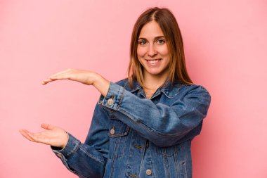 Pembe arka planda izole edilmiş genç beyaz bir kadın işaret parmaklarıyla küçük bir şey tutuyor, gülümsüyor ve kendinden emin..