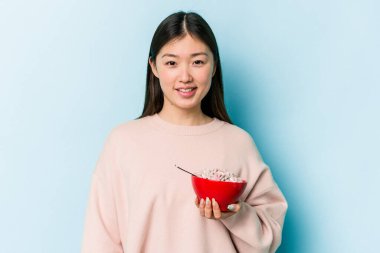 Mavi arka planda bir kase mısır gevreği tutan genç Asyalı kadın mutlu, gülümseyen ve neşeli..