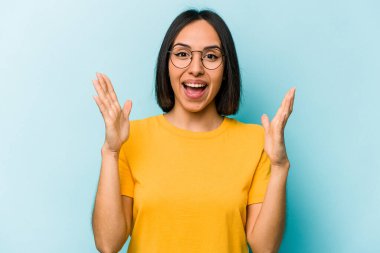 Genç İspanyol kadın mavi arka planda izole edilmiş zaferi ya da başarıyı kutluyor, şaşırmış ve şok olmuş..