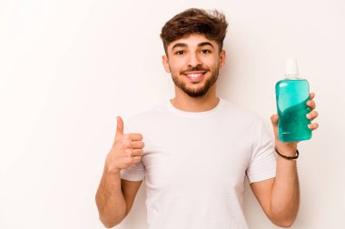 Beyaz arka planda ağız gargarası tutan genç İspanyol adam gülümsüyor ve başparmağını kaldırıyor.
