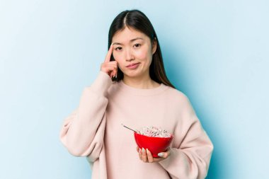 Genç Asyalı bir kadın elinde bir kase mısır gevreği ile mavi arka planda izole edilmiş bir şekilde parmağıyla tapınağı işaret ediyor, düşünüyor ve bir göreve odaklanıyor..