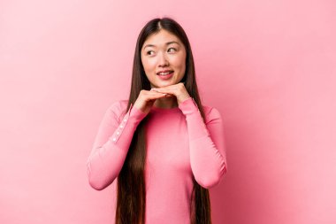 Pembe arka planda izole edilmiş genç Çinli kadın ellerini çenesinin altında tutuyor ve mutlu bir şekilde yan tarafa bakıyor..