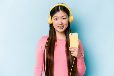 Genç Asyalı kadın mavi arka planda izole edilmiş müzik dinlerken gülüyor ve eğleniyor..