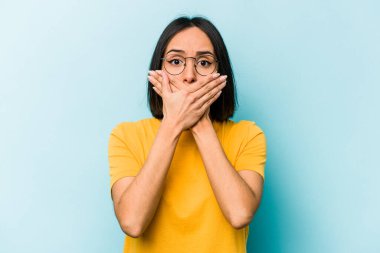 Mavi arka planda izole edilmiş genç İspanyol kadın elleriyle ağzını kapatırken şok oldu..
