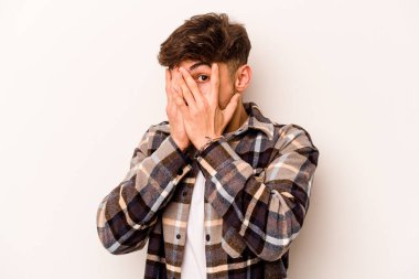 Beyaz arka planda izole edilmiş genç İspanyol adam parmaklarının arasından korkmuş ve gergin bir şekilde göz kırpıyor..