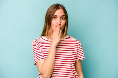 Mavi arka planda izole edilmiş genç beyaz kadın şok olmuş, ağzı ellerle kaplanmış, yeni bir şey keşfetmeye hevesli..