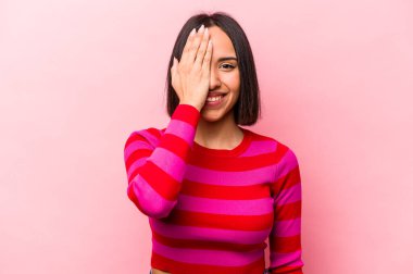 Pembe arka planda izole edilmiş genç İspanyol kadın yüzünün yarısını avuç içiyle kaplayarak eğleniyor..