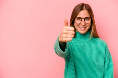 Pembe arka planda izole edilmiş genç beyaz kadın gülümsüyor ve başparmağını kaldırıyor.