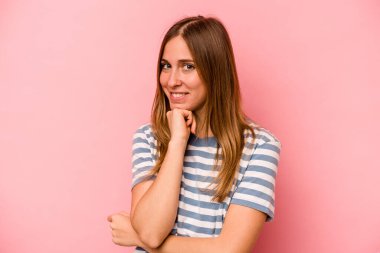 Pembe arka planda izole edilmiş genç beyaz kadın gülümsüyor ve kendinden emin, elleriyle çenesine dokunuyor..