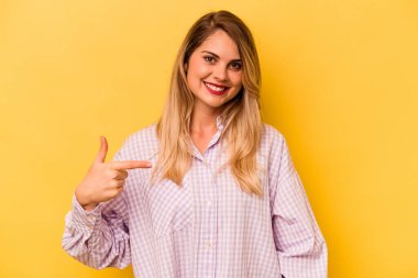 Sarı arka planda izole edilmiş genç beyaz bir kadın gömleğin fotokopi odasını elle işaret ediyor, gururlu ve kendinden emin.