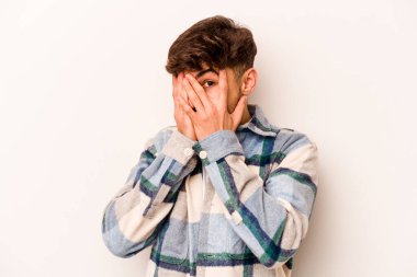 Beyaz arka planda izole edilmiş genç İspanyol adam parmaklarının arasından korkmuş ve gergin bir şekilde göz kırpıyor..