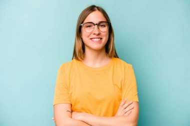 Mavi arka planda izole edilmiş genç beyaz kadın kendini güvende hissediyor, kararlılıkla kollarını kavuşturuyor..