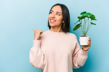 Mavi arka planda bir bitkiyi tutan genç İspanyol kadın başparmak uzakta, gülerek ve kaygısızca.