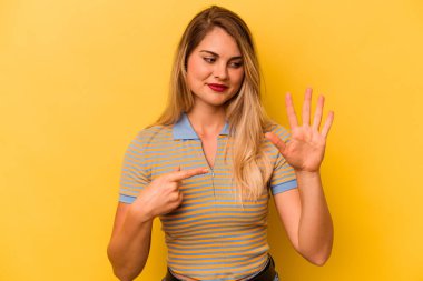 Sarı arka planda izole edilmiş genç beyaz kadın gülümseyerek beş numarayı parmaklarıyla gösteriyor..