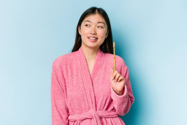 Bornoz giymiş genç Asyalı bir kadın pembe arka planda izole edilmiş dişlerini fırçalıyor. Gülümsüyor, neşeli ve keyifli görünüyor..