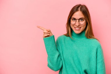 Pembe arka planda izole edilmiş genç beyaz kadın işaret parmağıyla işaret ederek gülümsüyor..