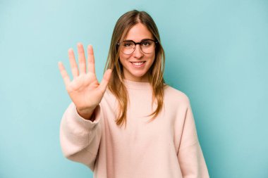 Mavi arka planda izole edilmiş genç beyaz kadın gülümseyerek beş numarayı parmaklarıyla gösteriyor..