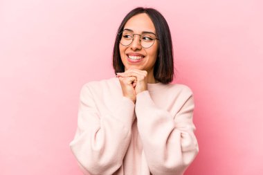 Pembe arka planda izole edilmiş genç İspanyol kadın ellerini çenesinin altında tutuyor ve mutlu bir şekilde yan tarafa bakıyor..