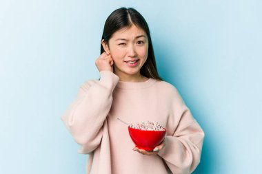 Genç Asyalı bir kadın elinde bir kase mısır gevreği tutuyor mavi arka planda izole edilmiş kulakları elleriyle kapatıyor..