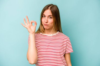 Kafkasyalı genç bir kadın mavi arka planda izole edilmiş bir şekilde bir göz kırpıyor ve elleriyle iyi bir jest yapıyor..