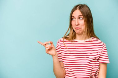Mavi arka planda izole edilmiş genç beyaz kadın gülümsüyor ve boş alanda bir şeyler gösteriyor..