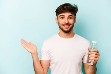 Young hispanic man holding a bottle of water isolated on white background showing a copy space on a palm and holding another hand on waist.