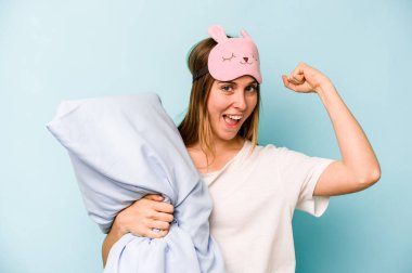Mavi arka planda izole edilmiş pijama giyen beyaz bir kadın zafer kazandıktan sonra yumruğunu kaldırıyor..