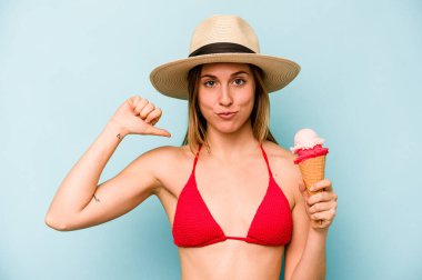 Bikini giyen ve elinde dondurma tutan beyaz bir kadın mavi arka planda kendini gururlu ve kendinden emin hissediyor..