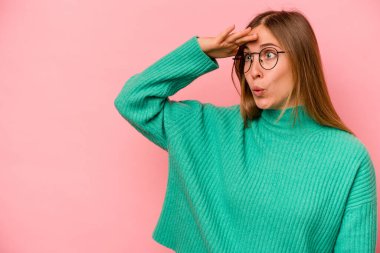 Pembe arka planda izole edilmiş genç beyaz kadın uzaklara bakıyor ve elini alnına koyuyor..