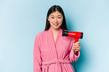 Bornoz giyen ve elinde saç kurutma makinesi tutan genç Asyalı kadın pembe arka planda izole edilmiş mutlu, güler yüzlü ve neşeli..