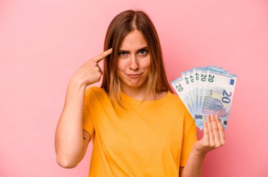 Elinde pembe arka planda izole edilmiş banknotlar tutan beyaz bir kadın işaret parmağıyla yaptığı hayal kırıklığı jestini gösteriyor..