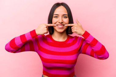 Pembe arka planda izole edilmiş genç İspanyol kadın gülümsüyor, parmaklarını ağıza doğrultuyor..