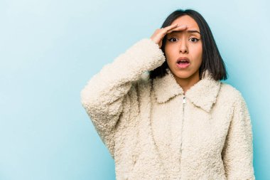 Mavi arka planda izole edilmiş genç İspanyol kadın uzaklara bakıyor ve elini alnına koyuyor..