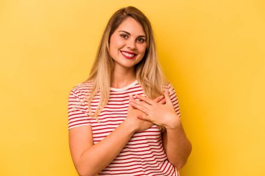 Sarı arka planda izole edilmiş genç beyaz kadın gülüyor, elini yüreğine koyuyor, mutluluk kavramını koruyor..