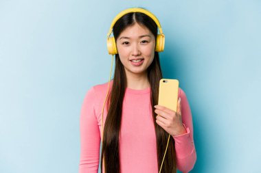 Mavi arka planda müzik dinleyen genç Asyalı kadın mutlu, güler yüzlü ve neşeli..