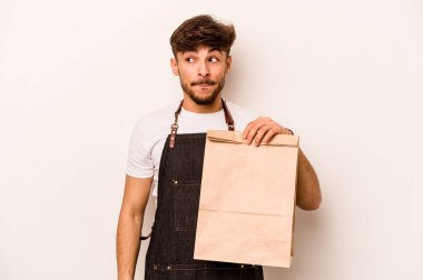 Beyaz arka planda izole edilmiş bir çanta taşıyan genç İspanyol tezgahtar kafası karışmış ve kendinden emin değil..