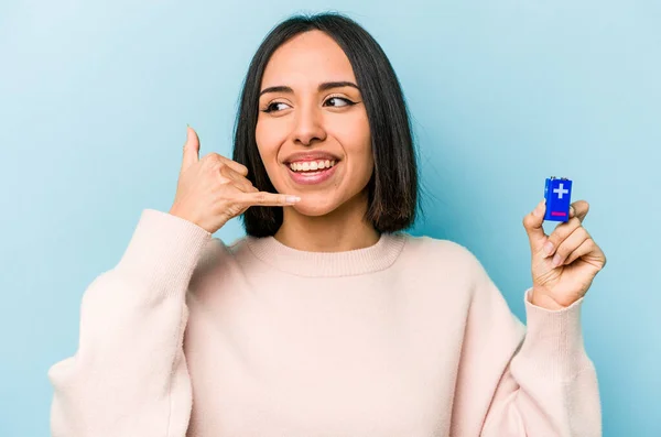 青の背景にバッテリーを持っている若いヒスパニック系の女性は指で携帯電話の呼び出しジェスチャーを示しています — ストック写真
